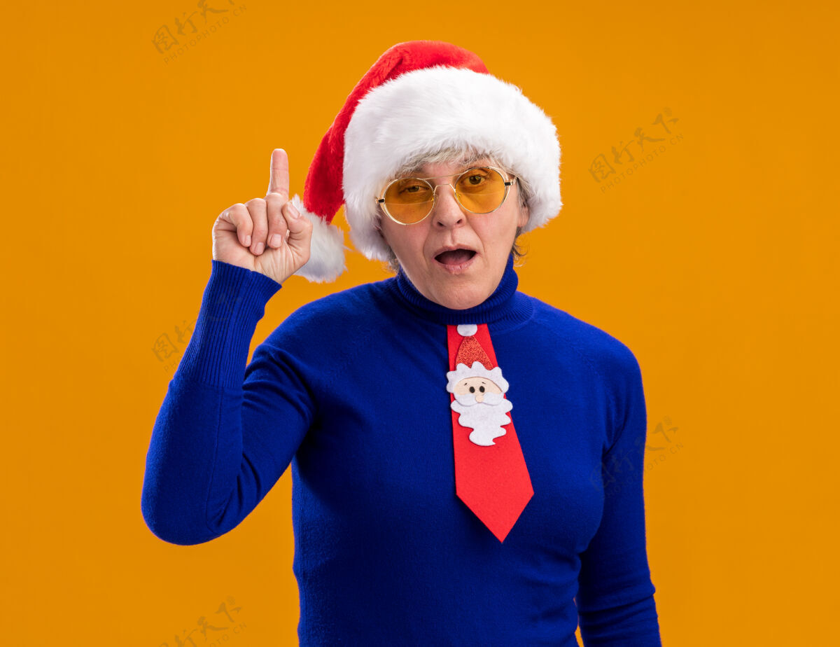 眼镜一位戴着太阳眼镜 戴着圣诞帽 系着圣诞领带的惊讶的老妇人 在橙色背景上孤立地指向上 留有复制空间指着帽子惊讶