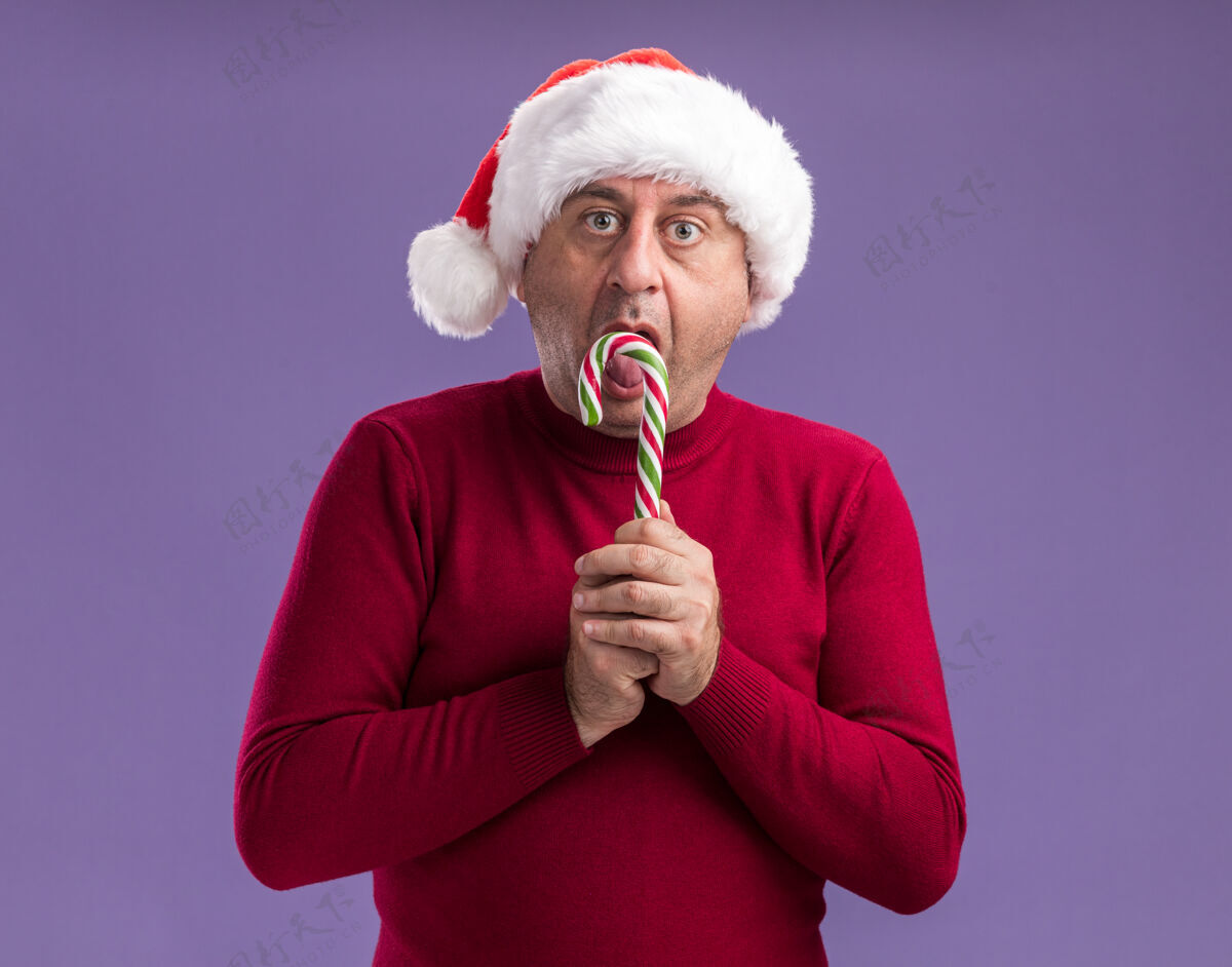 站着中年男子戴着圣诞老人帽手持糖果手杖站在紫色背景下惊讶地看着相机糖果惊讶中间