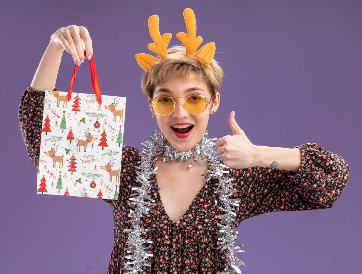 圣诞节令人印象深刻的年轻漂亮女孩戴着驯鹿鹿角头带 脖子上戴着金箔花环 戴着眼镜 手里拿着圣诞礼品袋 在紫色的墙上孤立地竖起大拇指周围印象深刻脖子