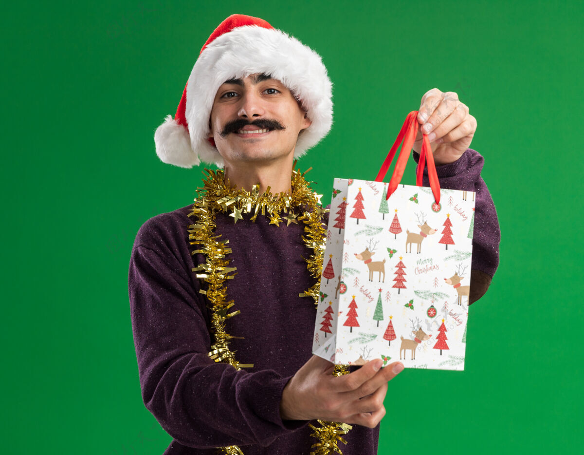 礼物快乐的小胡子男人戴着圣诞老人帽子 脖子上戴着金属丝 手里拿着圣诞礼物的纸袋 看着相机 微笑着自信地站在绿色的背景上年轻微笑金属片