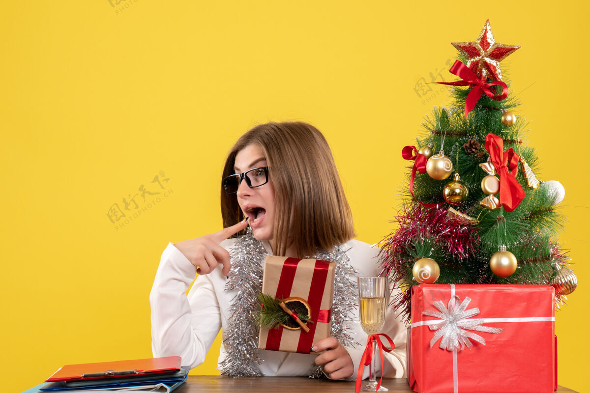 人正面图：女医生坐在桌子前 手里拿着黄色背景的礼物 还有圣诞树和礼品盒年办公室漂亮