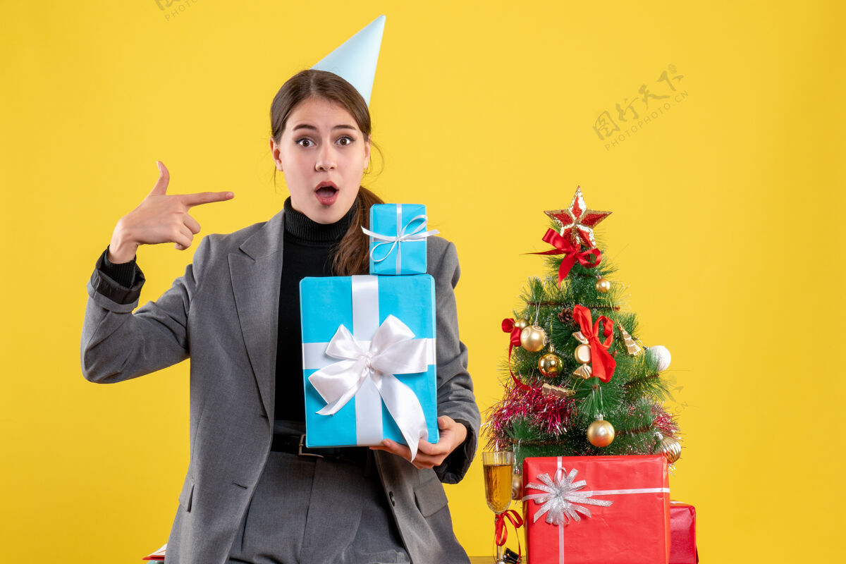 衣服前视图困惑的女孩与党帽举行圣诞礼物圣诞树附近的礼物鸡尾酒近女孩头饰