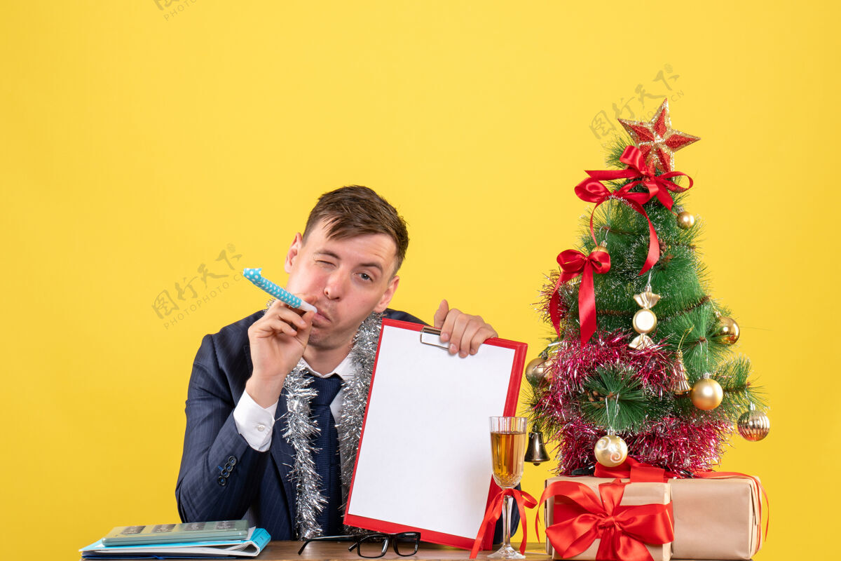 礼物商务人士使用noisemaker坐在圣诞树旁的桌子前 呈现黄色桌子树男人
