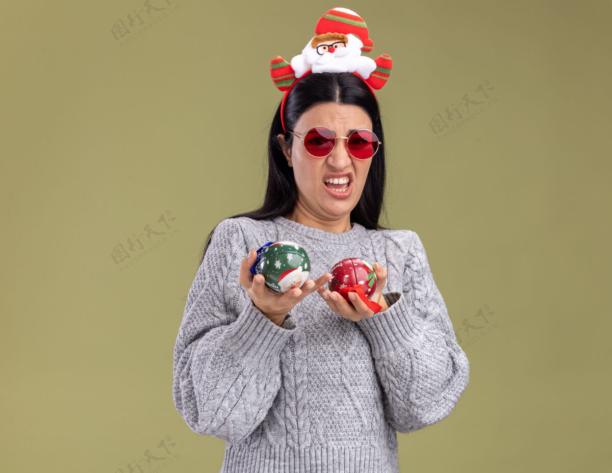 橄榄恼怒的年轻白人女孩戴着圣诞老人的头带和眼镜拿着圣诞饰品孤立在橄榄绿墙上的复制空间烦恼头带戴着