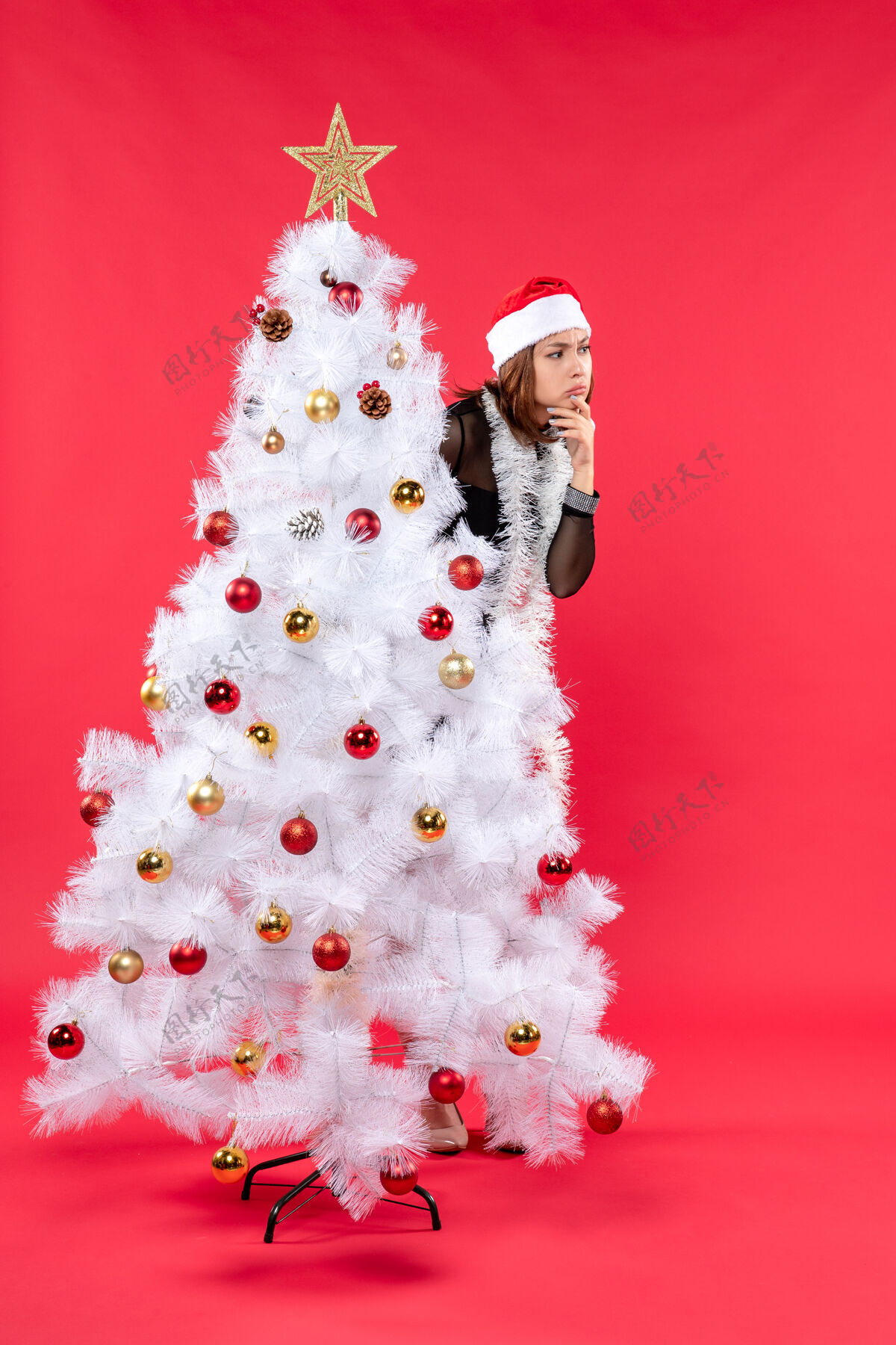 圣诞节圣诞心情与震惊年轻美丽的女士穿着黑色礼服和圣诞老人的帽子藏在新年树后庆祝新娘圣诞老人