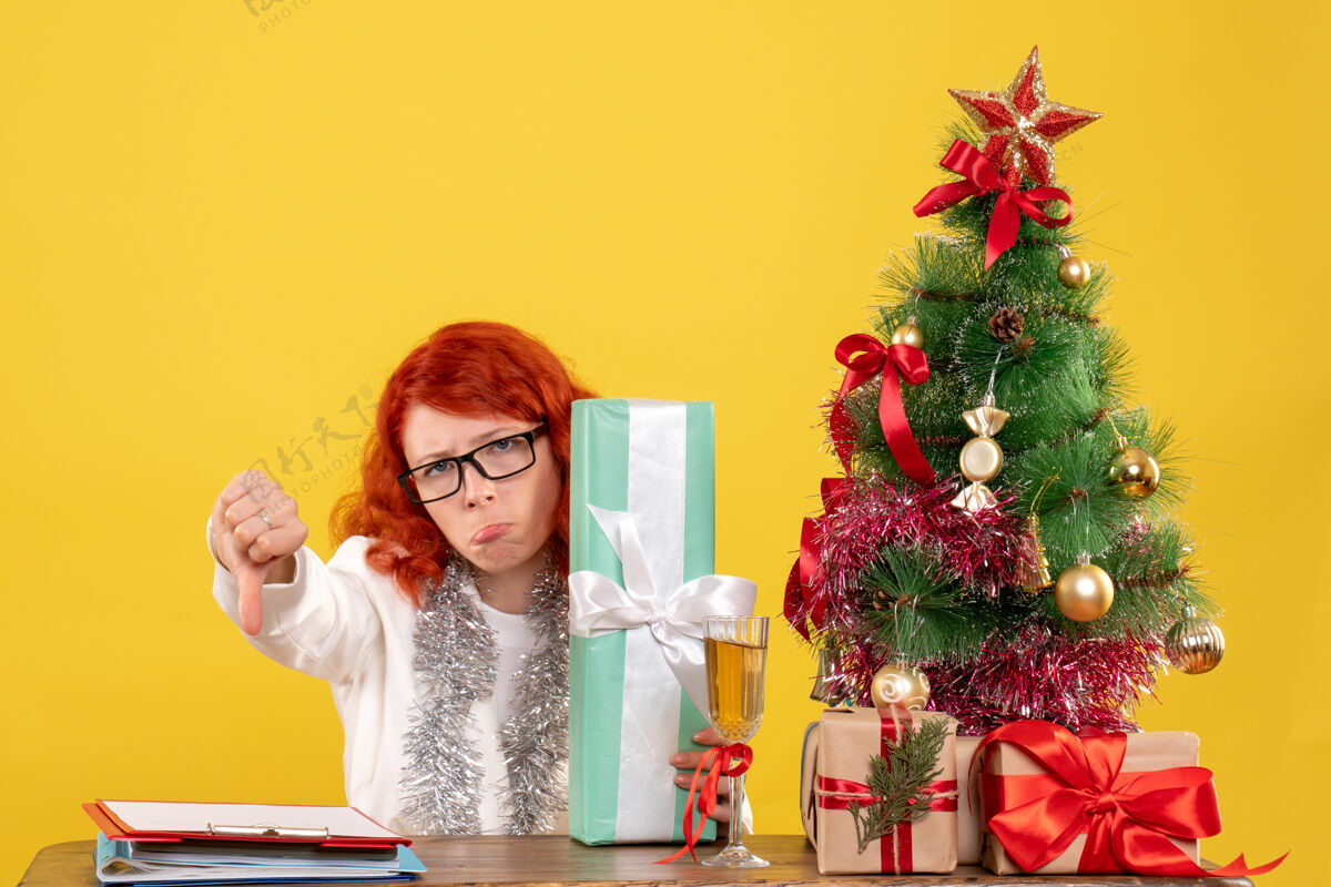 坐前视图：女医生拿着圣诞礼物和黄色背景上的圣诞树坐着医生庆祝前面