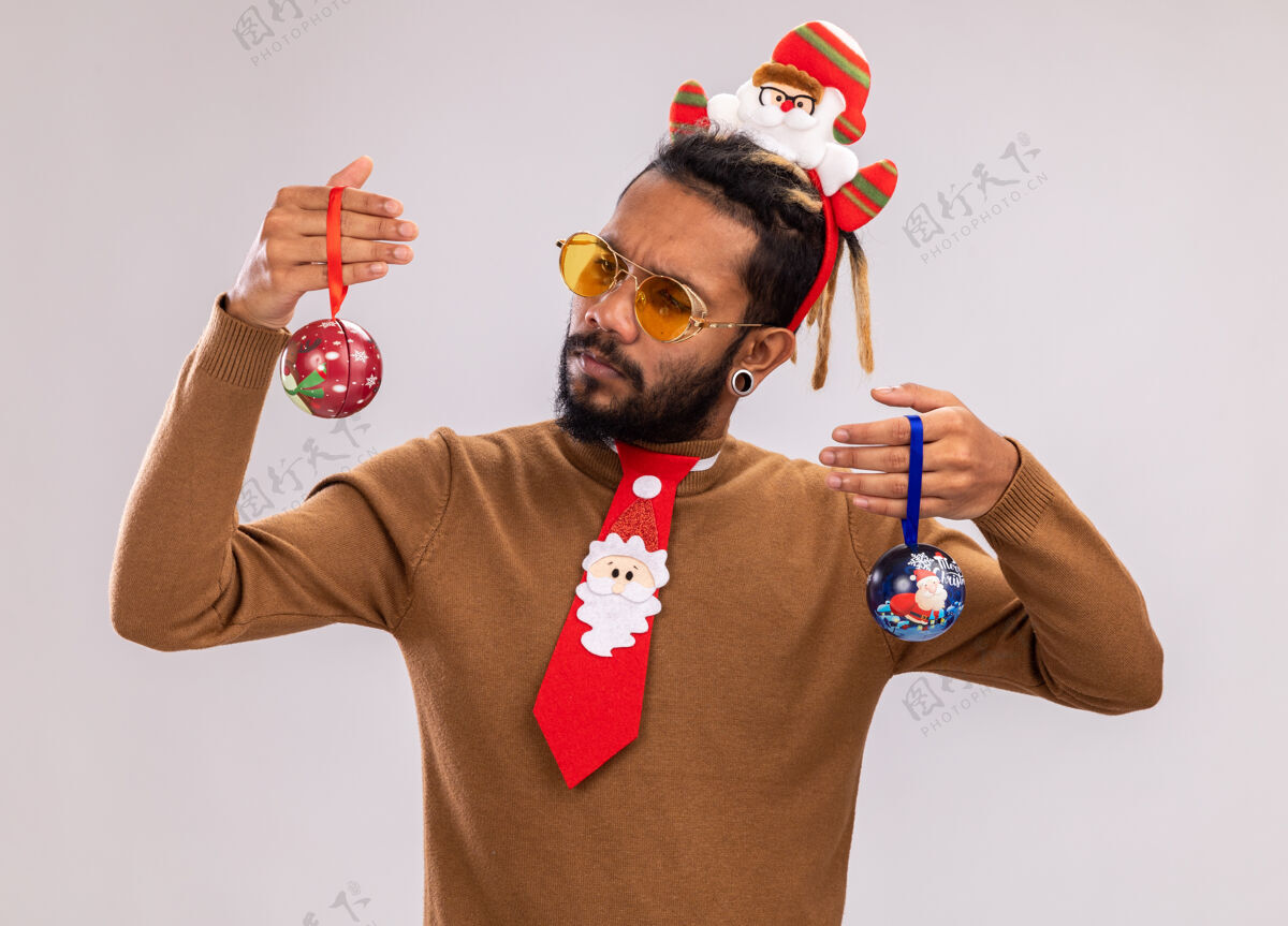困惑一个非洲裔美国人 穿着棕色毛衣 头戴圣诞老人圈 打着有趣的红色领带 手里拿着圣诞球 站在白色的墙上 看起来很困惑 试图做出选择头边缘男人