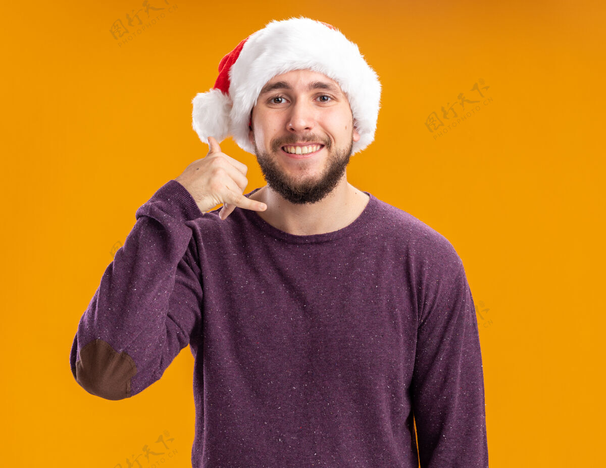 圣诞节穿着紫色毛衣 戴着圣诞帽的年轻人站在橘色的墙上 愉快而积极地向我打招呼积极毛衣圣诞老人