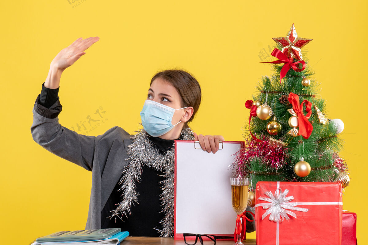 圣诞节正面图：戴着医用面罩的坚强年轻女孩坐在桌旁举手 圣诞树和鸡尾酒礼物礼物年轻坐着