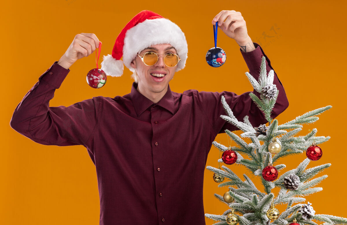 圣诞节快乐的年轻金发男子戴着圣诞帽和眼镜站在装饰过的圣诞树旁 手里拿着圣诞饰品 头被隔离在橙色的墙上圣诞饰品圣诞老人快乐