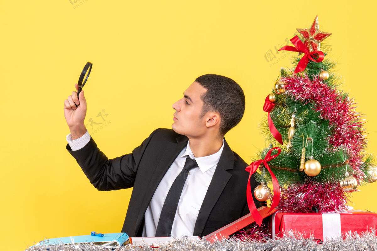 行政正面图：年轻人手里拿着光学lupa坐在桌旁圣诞树和礼物男人礼物Lupa