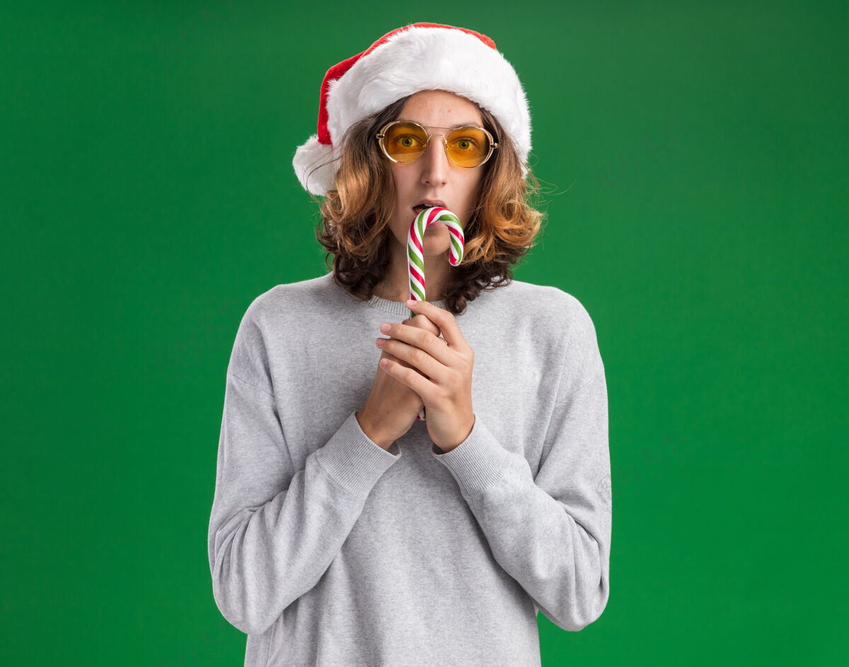 手杖戴着圣诞老人帽戴着黄色眼镜拿着糖果手杖的年轻人站在绿色背景下惊讶地看着相机糖果帽子抱着