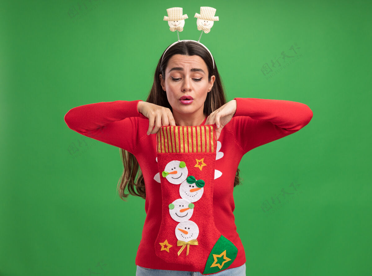 年轻人令人印象深刻的年轻美丽的女孩穿着圣诞毛衣与圣诞发箍举行 并期待着圣诞袜子隔离在绿色的墙壁袜子圣诞节毛衣