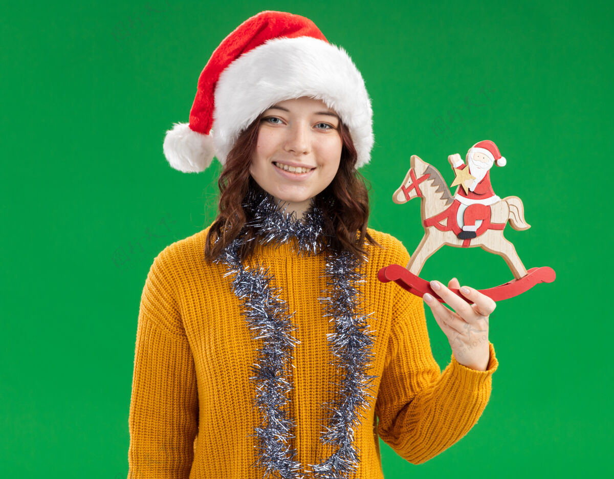 马微笑着的年轻斯拉夫女孩 戴着圣诞帽 脖子上戴着花环 把圣诞老人抱在摇摇木马上 绿色背景上有复制空间抱着帽子斯拉夫