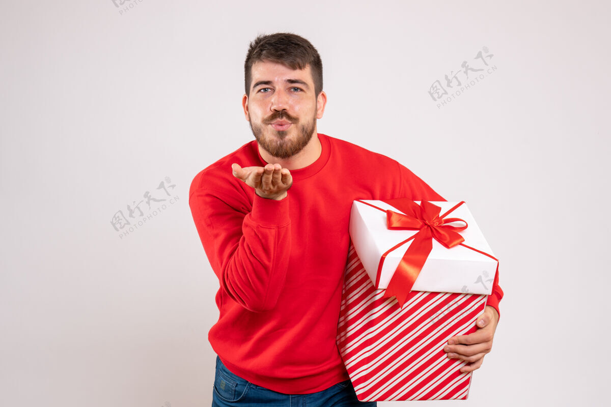人身穿红衫的年轻人拿着盒子里的圣诞礼物 在白墙上送吻吻人节日