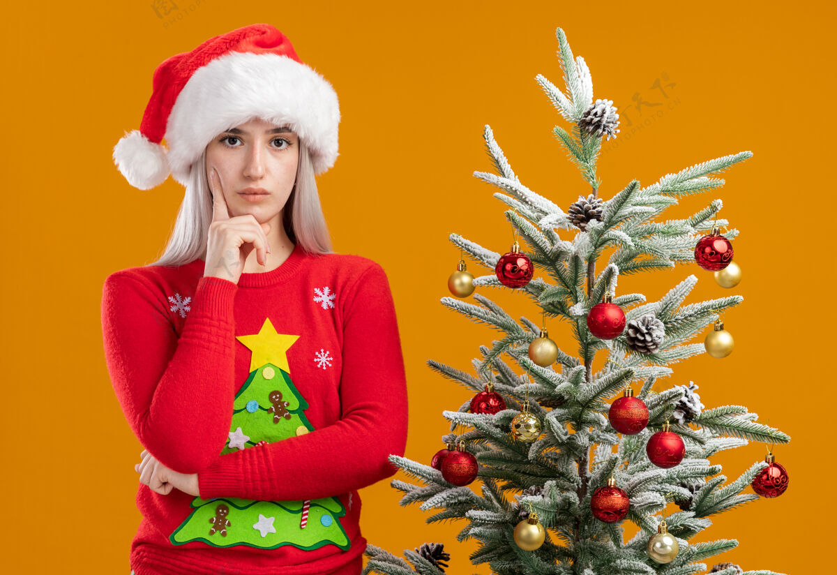 圣诞老人穿着圣诞毛衣 戴圣诞帽的金发女郎 手放在下巴上看着摄像机 严肃的脸站在橙色背景下的圣诞树旁思考毛衣圣诞金发