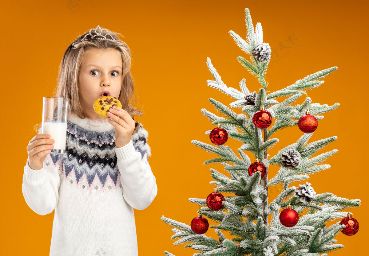 花环惊讶的小女孩站在圣诞树旁 戴着头饰 脖子上戴着花环 手里拿着一杯牛奶 尝试着橙色背景下的饼干饼干圣诞节手捧