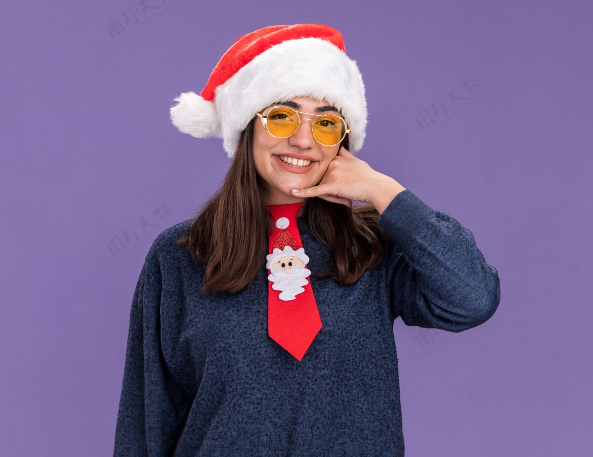 年戴着太阳眼镜 戴着圣诞帽 系着圣诞领带 微笑着的年轻白种人女孩在紫色背景上孤立地打着“呼叫我”的手势 并留有复制空间领带圣诞老人手势