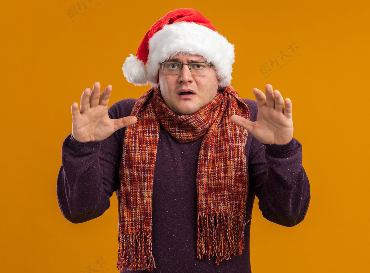 害怕一个戴着眼镜 戴着圣诞帽 脖子上围着围巾的惊恐的成年人看着摄像机 手举在空气中 在橙色背景下被隔离脖子眼镜圣诞老人
