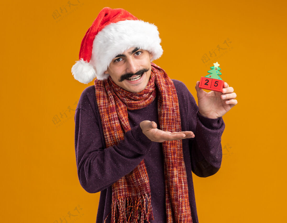 圣诞节快乐的胡子男人戴着圣诞老人帽 脖子上围着暖和的围巾 手里拿着日期为25的玩具立方体 手举着微笑站在橙色的背景上举行微笑周围
