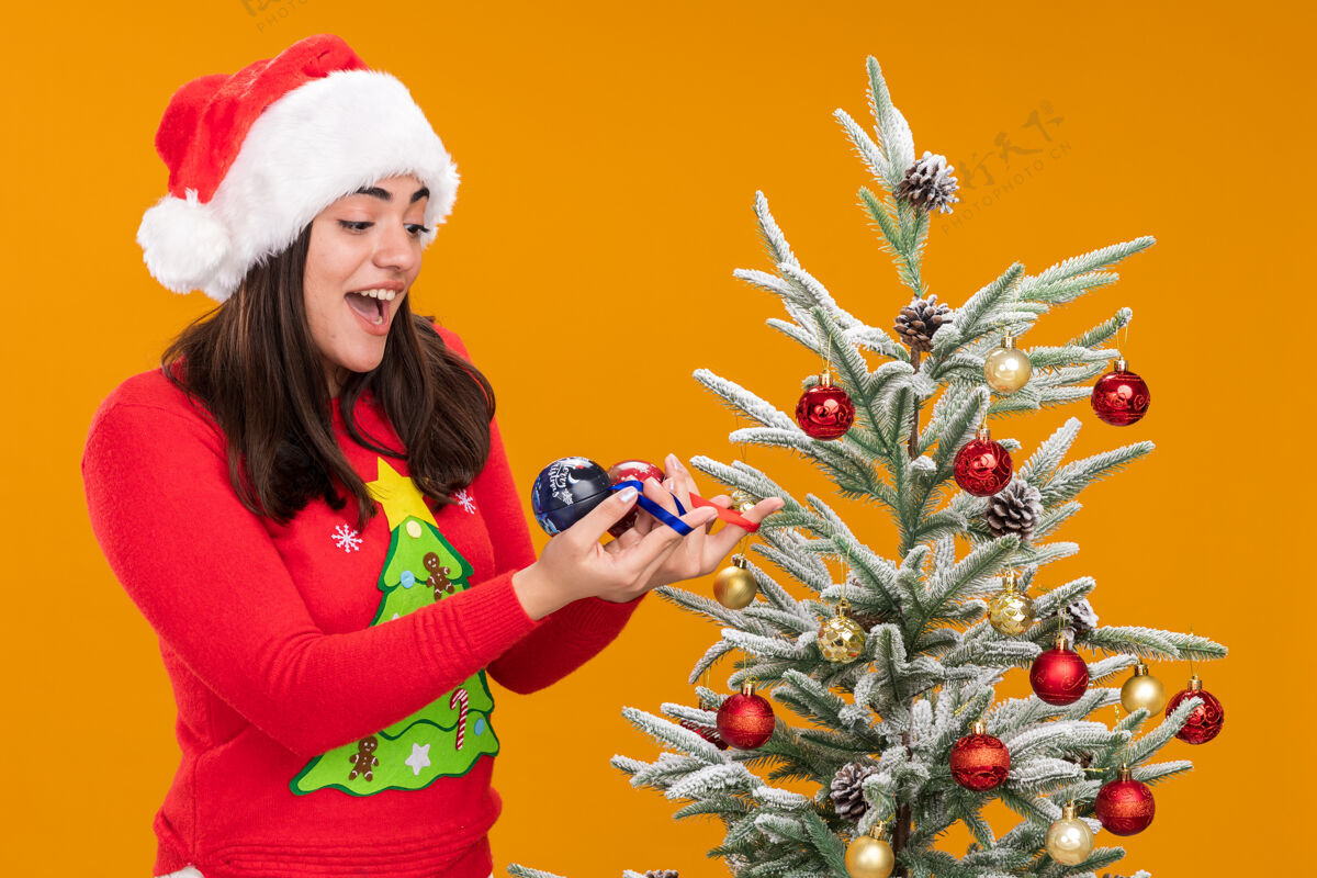 圣诞惊讶的年轻白人女孩拿着圣诞帽 看着玻璃球装饰站在旁边的圣诞树橙色背景上孤立的复制空间橙色球圣诞树