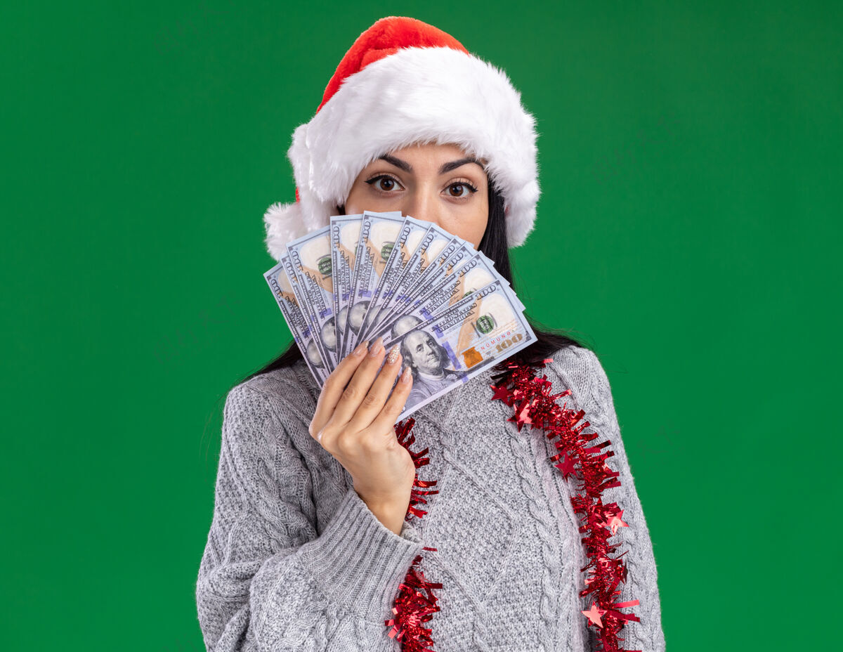 女孩年轻的高加索女孩戴着圣诞帽 脖子上戴着金箔花环 手里拿着钱 从后面看着隔离在绿色背景上的相机金属片花环拿着