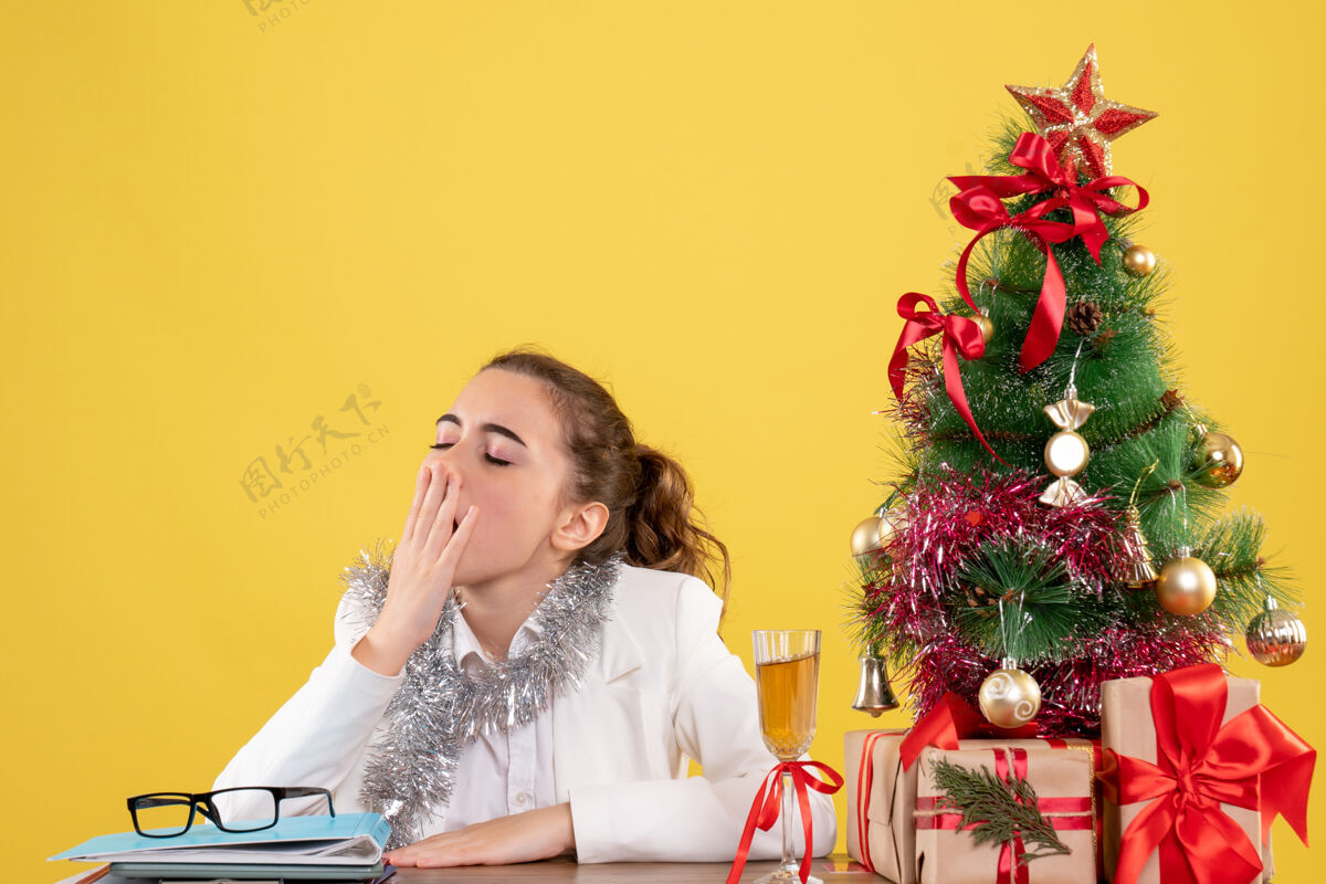 快乐正面图：女医生坐在桌子后面 打着哈欠 背景是黄色的圣诞树和礼盒女医生漂亮前面