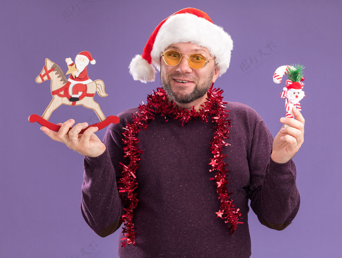 穿着快乐的中年男子戴着圣诞帽 脖子上戴着金箔花环 戴着眼镜 手里拿着糖果手杖饰品 圣诞老人骑着摇马 雕像孤立在紫色的墙上男人金属片脖子