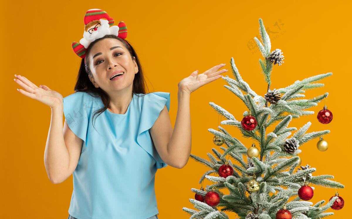 橘子身穿蓝色上衣的年轻女子头戴滑稽的圣诞环 看着摄像机 困惑地微笑着 双臂举在橙色背景下的圣诞树旁手臂边缘升起
