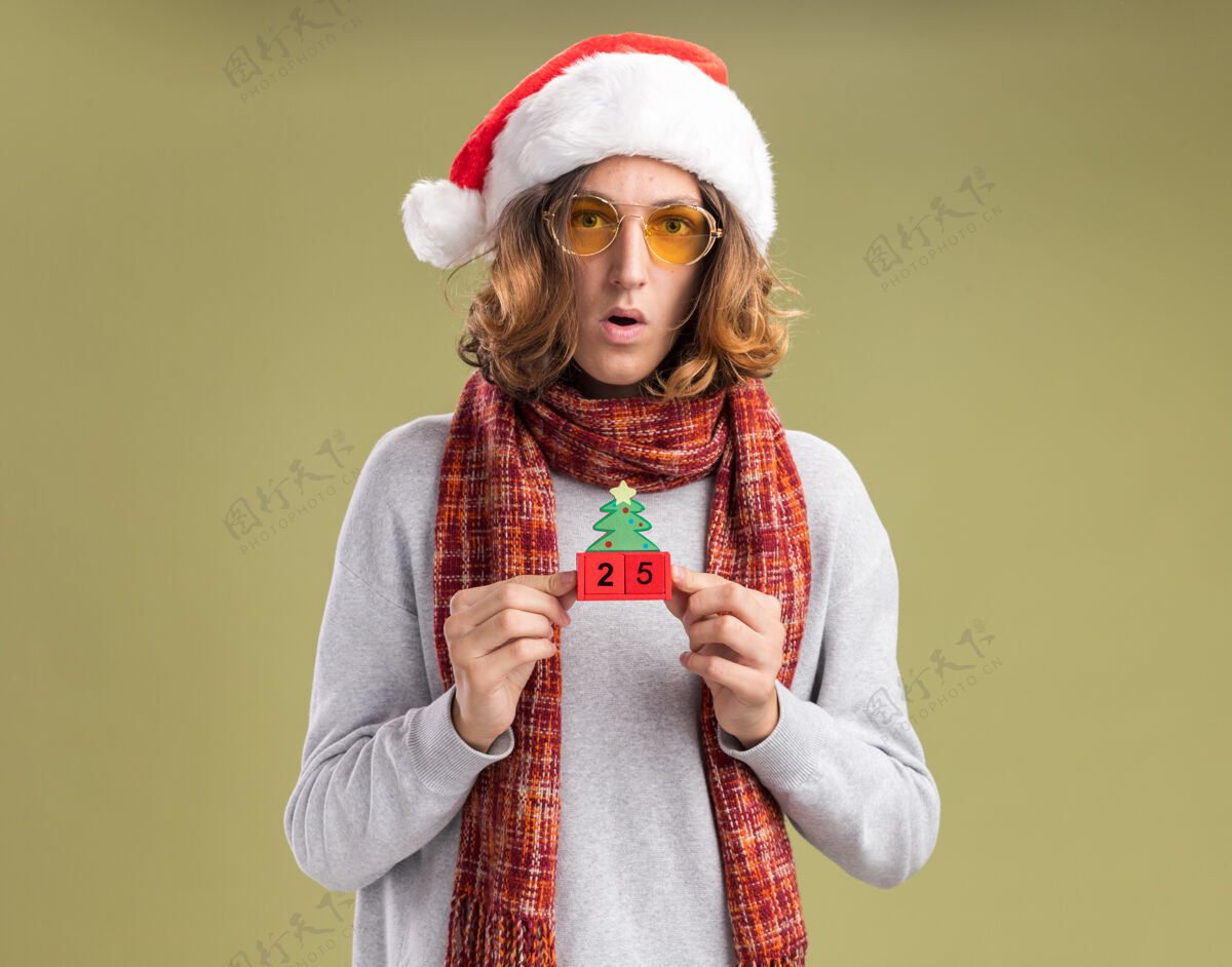 眼镜戴着圣诞老人帽 戴着黄色眼镜 脖子上围着暖和的围巾的年轻人 手里拿着和25岁约会的玩具立方体 站在绿色背景下 困惑而惊讶地看着摄像机相机看背景
