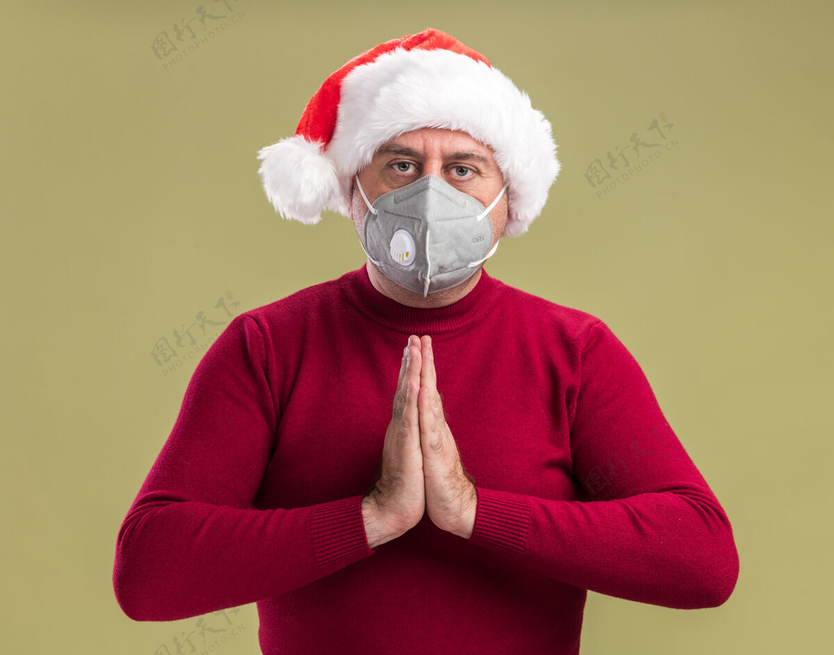 圣诞老人中年男子戴着圣诞老人帽戴着护面面具手拉着手像祈祷一样严肃地站在绿色的背景上一起脸年龄
