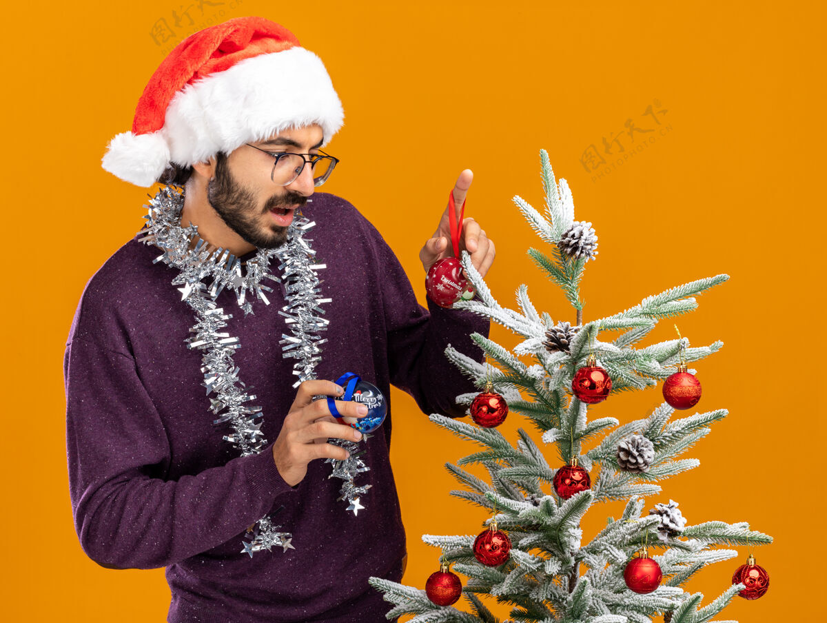 帽子想着站在圣诞树旁的年轻帅哥 戴着圣诞帽 脖子上戴着花环 手里拿着橙色背景上孤立的圣诞球小伙子花环思想