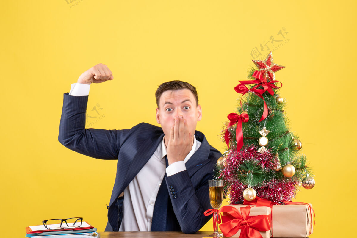 坐着令人惊讶的男子显示肌肉坐在圣诞树附近的桌子上 黄色墙上的礼物前视图展示男人西装