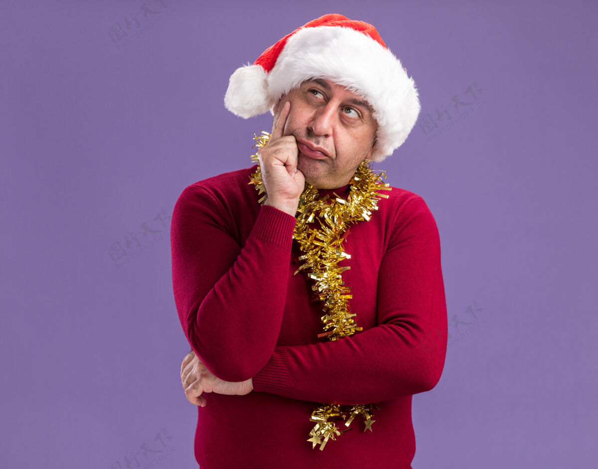 站立中年男子戴着圣诞老人的帽子 脖子上挂着金属丝 站在紫色的背景上 困惑地抬头看圣诞老人年龄金属片