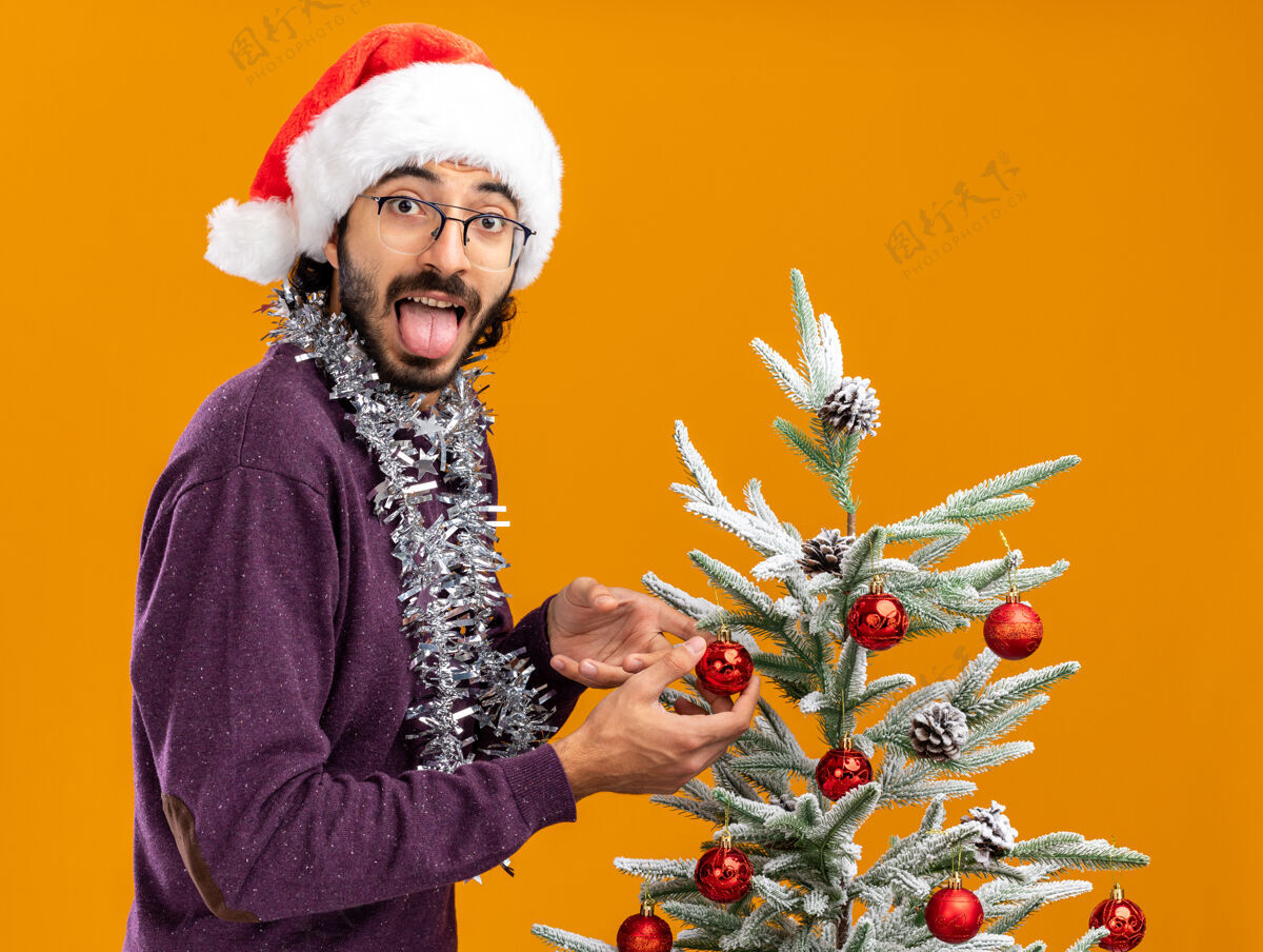 展示快乐的年轻帅哥站在圣诞树旁 戴着圣诞帽 脖子上戴着花环 舌头孤立在橙色的墙上就在附近站着帽子