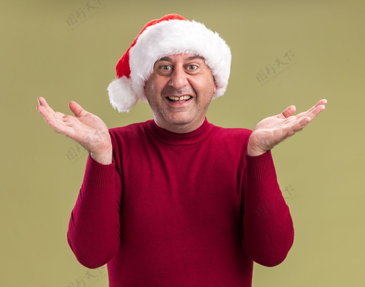 胳膊中年男子戴着圣诞老人的帽子 看着镜头 高兴而兴奋地微笑着 双臂高举 站在绿色的背景上年龄微笑圣诞节