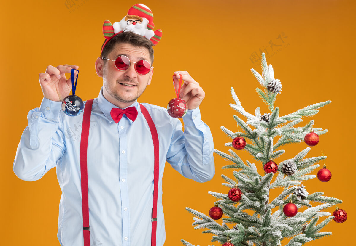 眼镜戴着吊带领结的年轻人站在圣诞树旁边 戴着圣诞老人和红眼镜 看起来很困惑 拿着玩具在橙色的背景下为圣诞树吊带轮辋领带