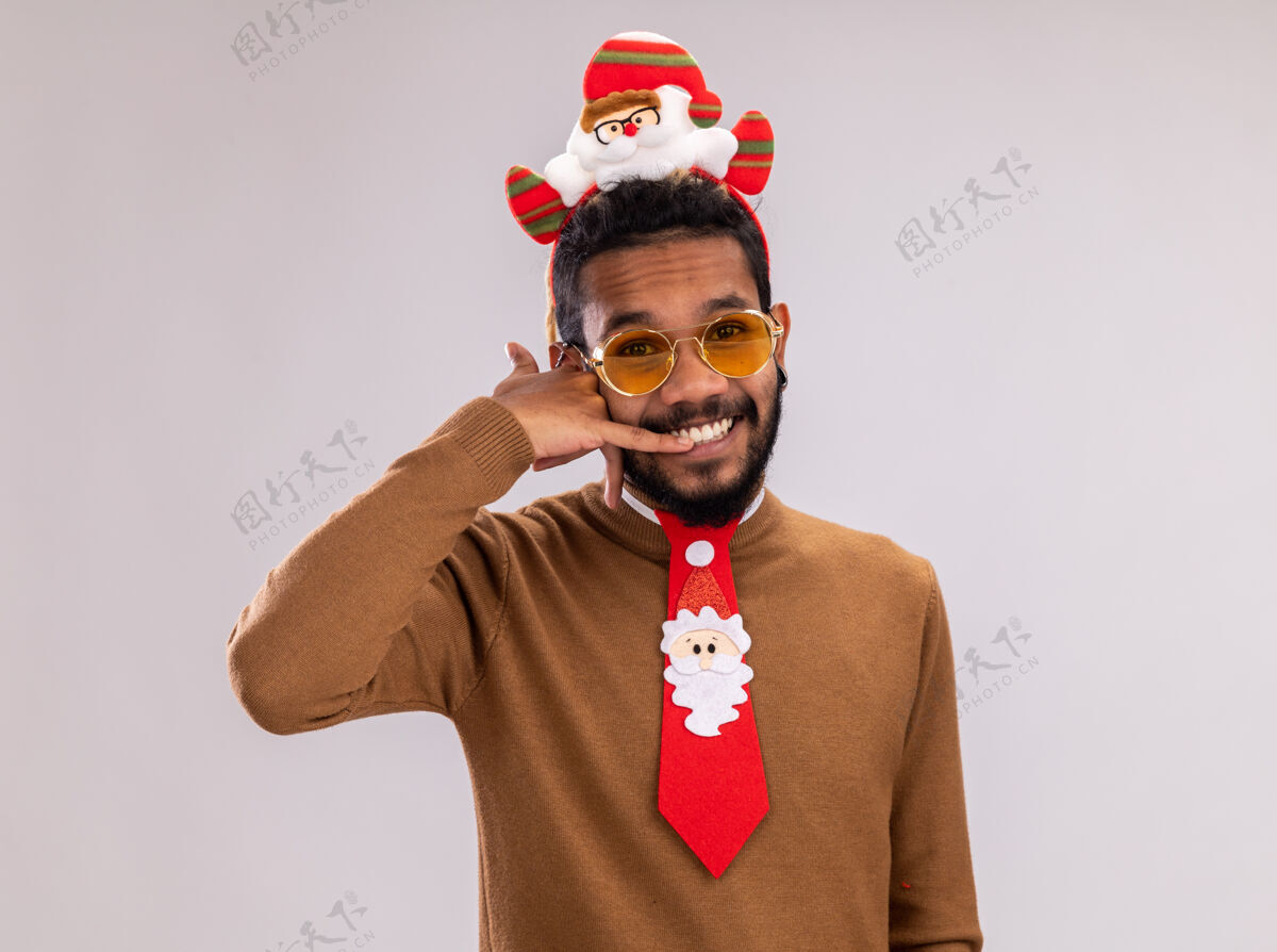 头非洲裔美国人 身穿棕色毛衣 头戴圣诞老人耳环 打着有趣的红色领带 站在白色背景上愉快地做着“叫我”的手势高兴手势边缘