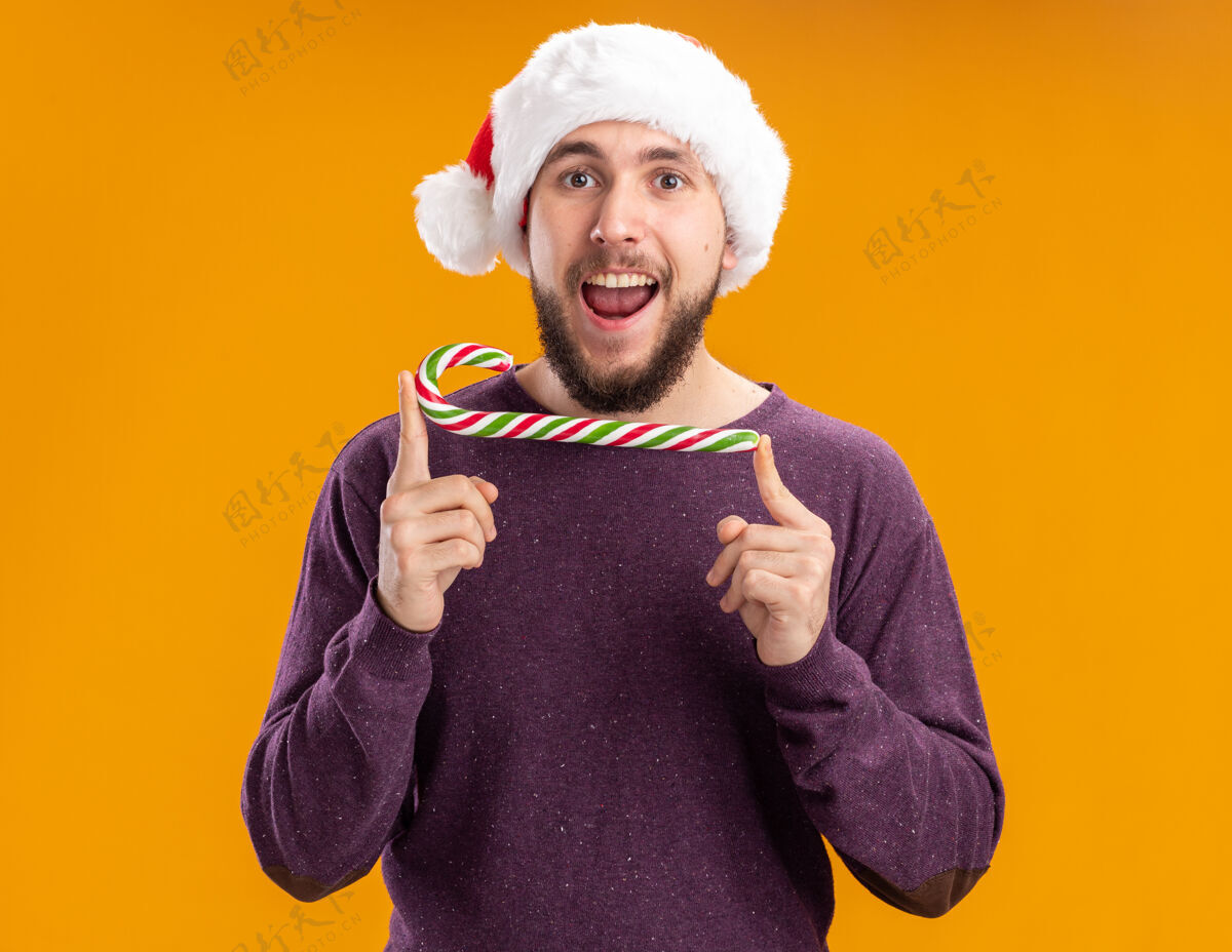 年轻人身穿紫色毛衣 戴圣诞帽的年轻人手持糖果手杖 站在橙色背景下 开心地看着镜头脸糖果拿着