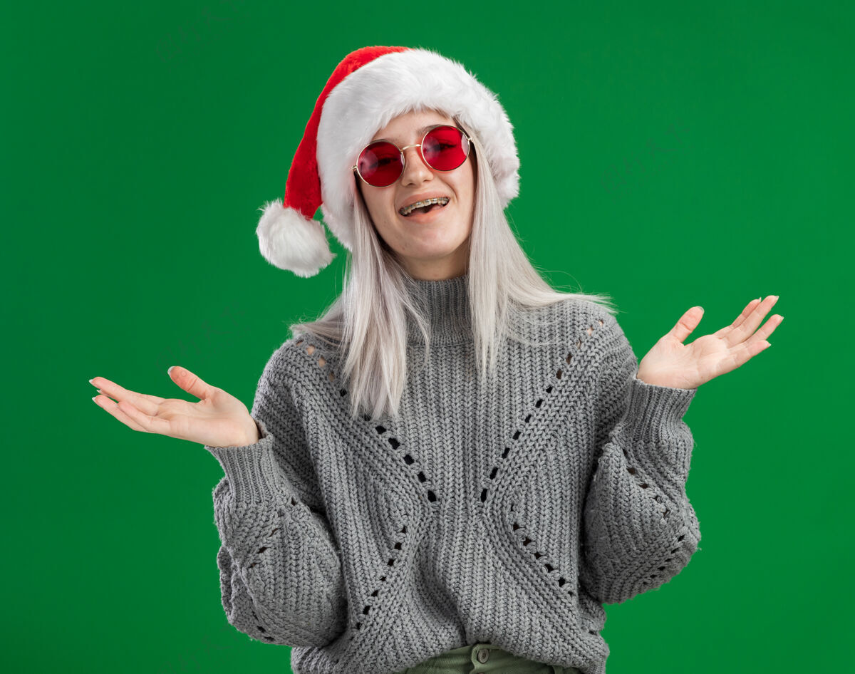 年轻年轻的金发女郎穿着冬衣 戴着圣诞帽 戴着红眼镜 看着镜头 快乐而积极地微笑着 欢快地站在绿色的背景下微笑帽子圣诞老人