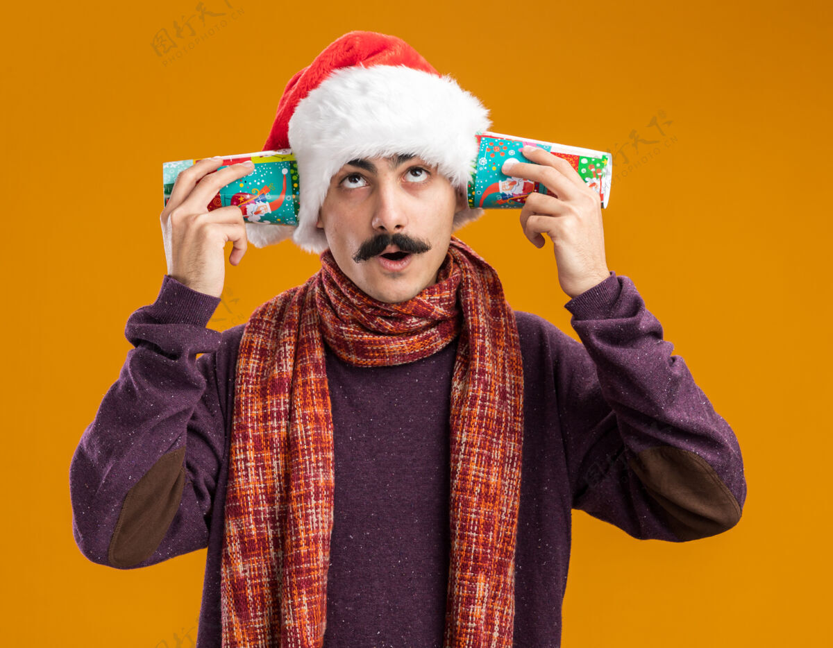 站着一个留着胡子的男人戴着圣诞老人帽 脖子上围着暖和的围巾 耳朵上戴着五颜六色的纸杯 站在橘色的背景上困惑地仰望着耳朵抱着温暖