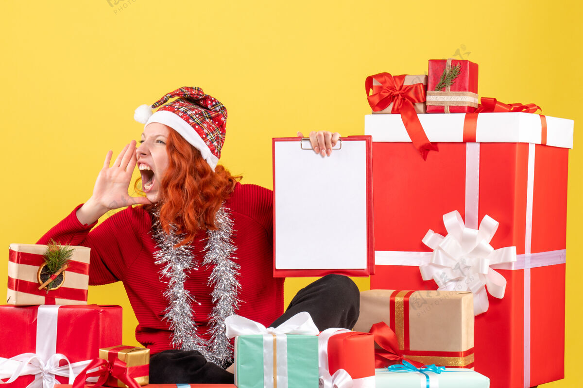 微笑坐在圣诞礼物周围的年轻女子的正面视图 黄色墙上有文件说明快乐前面礼物