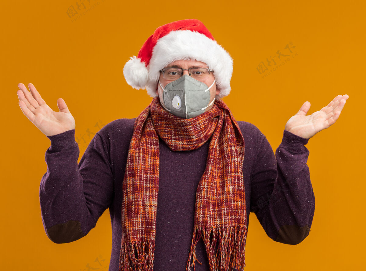 空笨手笨脚的成年男子戴着防护面具眼镜 戴着圣诞帽 脖子上围着围巾 看着摄像机 两手空空的 孤立在橙色背景上保护围巾帽子