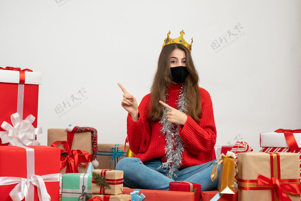 坐着穿着红色毛衣的年轻女孩手指着坐在那里的东西 礼物上戴着黑色的面具周围红色礼物