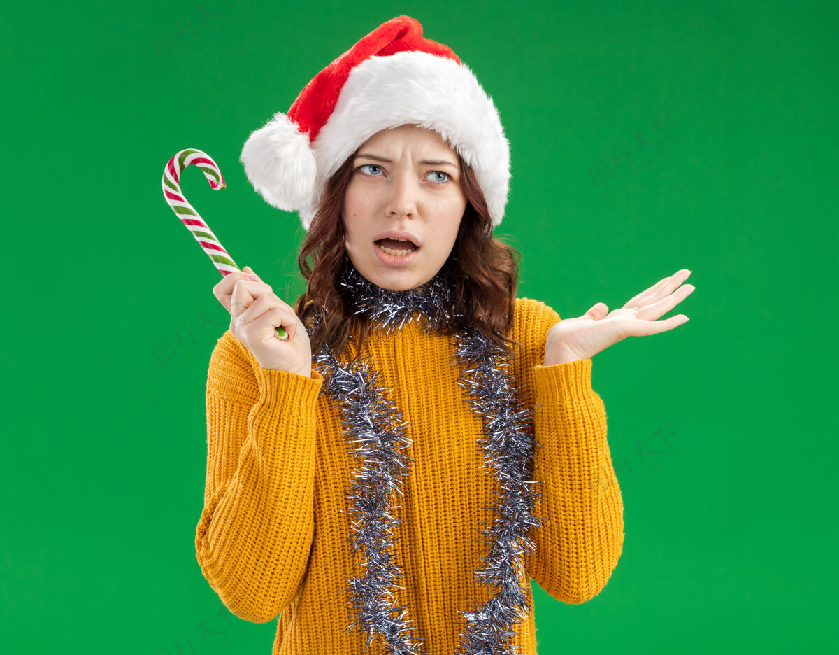 斯拉夫恼怒的斯拉夫女孩戴着圣诞帽 脖子上戴着花环 手里拿着糖果手杖 在绿色的背景上保持手的张开 还有复制空间花环帽子恼怒