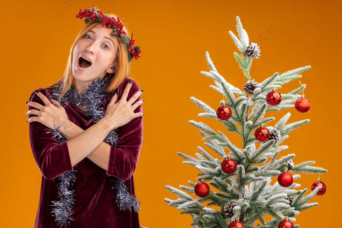 肩膀兴奋的年轻漂亮女孩站在圣诞树旁 身穿红色连衣裙 脖子上戴着花环 手放在肩膀上 孤立地站在橙色的墙上衣服圣诞节附近
