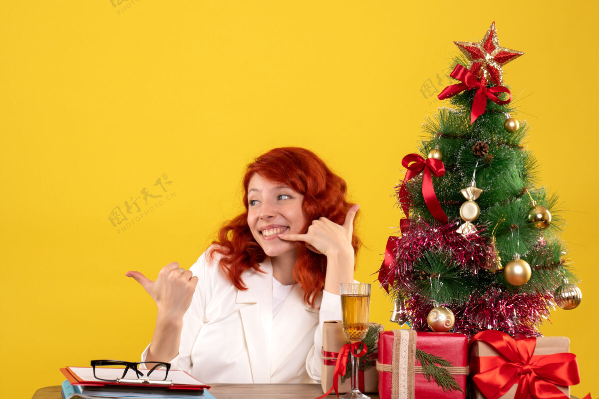 安排女工人坐在桌子后面 手里拿着圣诞礼物 树在黄色上微笑树情感工人