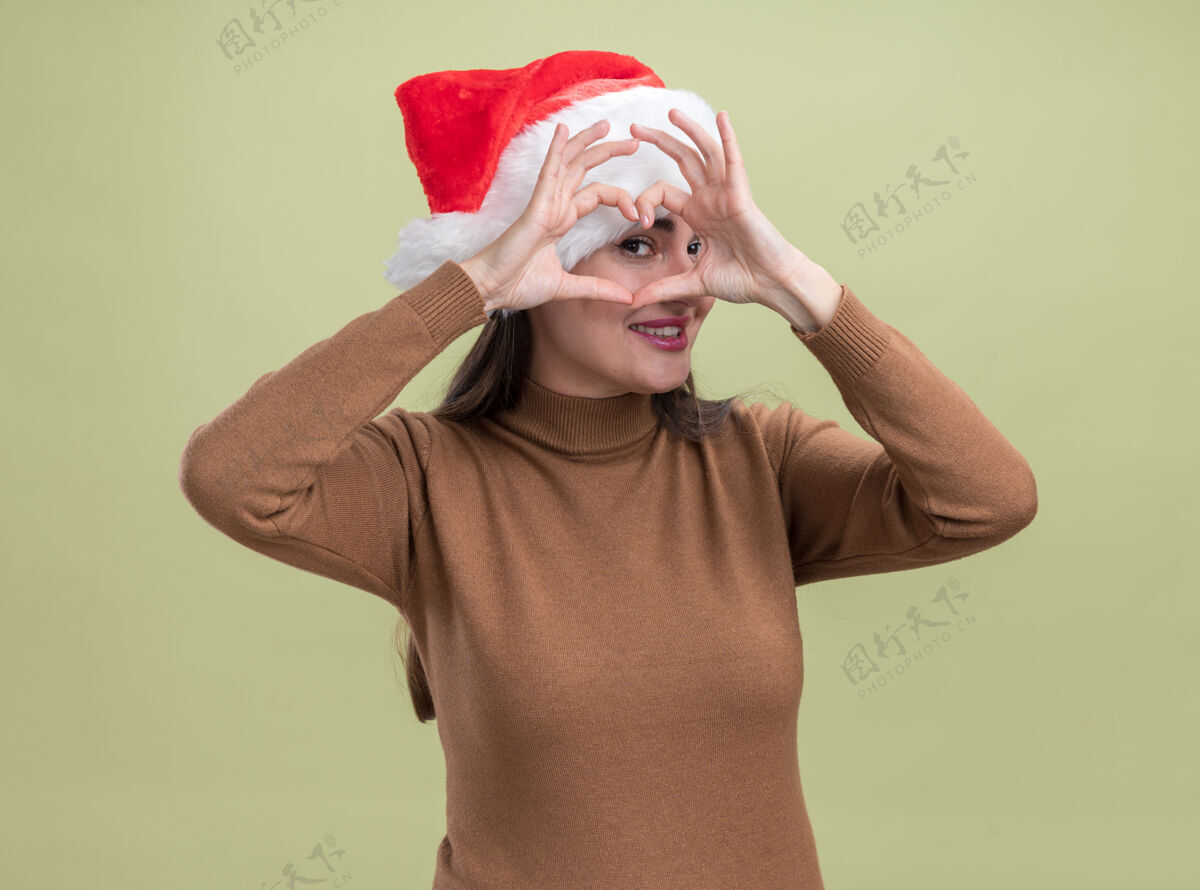 年轻微笑的年轻漂亮女孩戴着圣诞帽 在橄榄绿的背景上显示出孤立的心形手势帽子橄榄