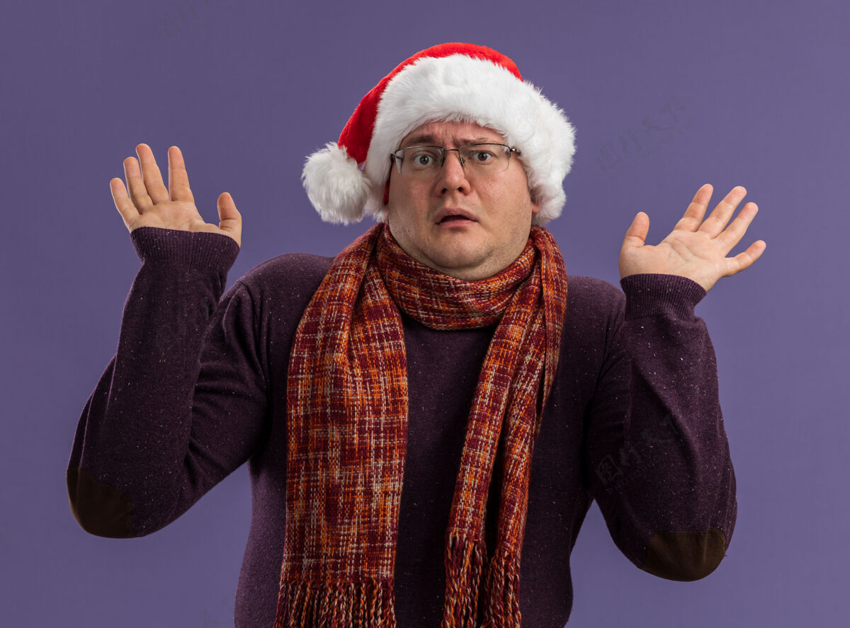 空笨手笨脚的成年男子戴着眼镜 戴着圣诞帽 脖子上围着围巾 看着镜头 紫色背景上孤立着空空的双手圣诞老人周围不知所措