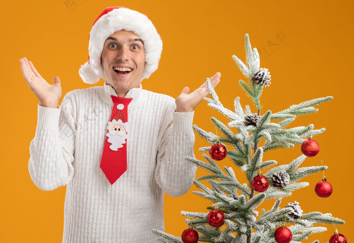 年轻令人印象深刻的年轻帅哥戴着圣诞帽 打着圣诞老人的领带 站在装饰好的圣诞树旁 两手空空的 孤零零地站在橙色的墙上帅气圣诞节秀着